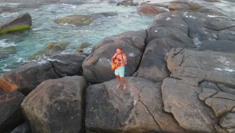 Aerial-top-down-shot-of-young-man-in-bathers-standing-on-rock-and-playing-guitar-in-front-of-ocean-and-beautiful-sunset,-Australia