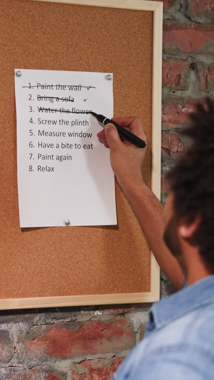African-American man crosses out accomplished to-do list points on cork board near brick wall closeup. Black worker checks plan of flat renovation