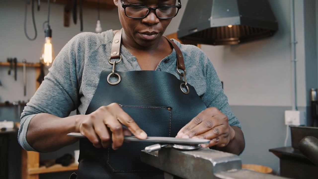 Donna con gli occhiali e grembiule di pelle che lima un disco di metallo in una morsa in officina, usando una lima a mano.
