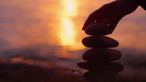 The Silhouette Of A Hand That Builds A Pyramid Of Stones Against The Background Of The Sea Where The