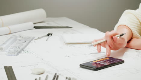 close-up of a person's hands working on architectural blueprints with drawing tools and a smartphone calculator