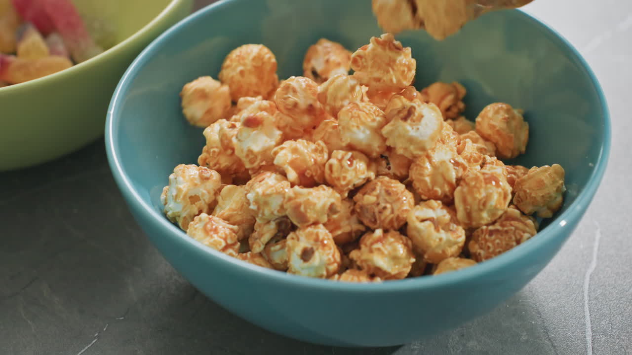 Close view of caramel flavored popcorn pouring from packet into blue bowl on dark surface, green bowl with colorful candy in background