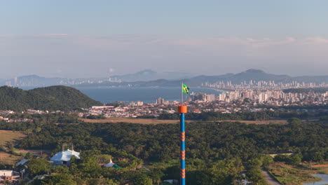 the brazilian flag waves proudly atop a tall ride structure at beto carrero world in penha, santa catarina, with the atlantic ocean and urban coastline visible in the background