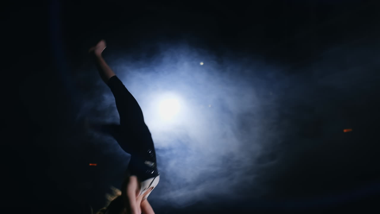 Young female gymnast runs up and performs flips forward and screw in the air in slow motion in gym on carpet in smoke.