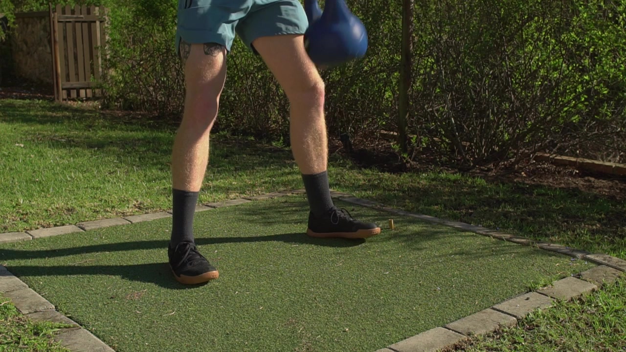 Jovem levanta um kettlebell azul do chão em um movimento de arremesso e desenvolvimento com um braço. Vista em close-up de baixo ângulo enquadrando suas pernas, pés e o kettlebell em um tapete de treino ao ar livre