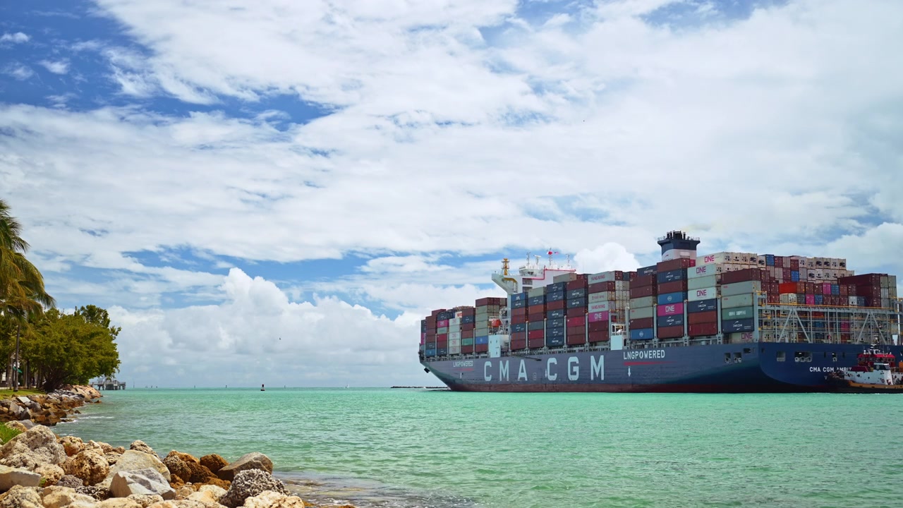 Miami, USA, 16 April 2026: Large CMA CGM container ship navigating near Miami coast. Side view of a blue cargo vessel loaded with colorful containers on turquoise ocean water under cloudy blue sky.