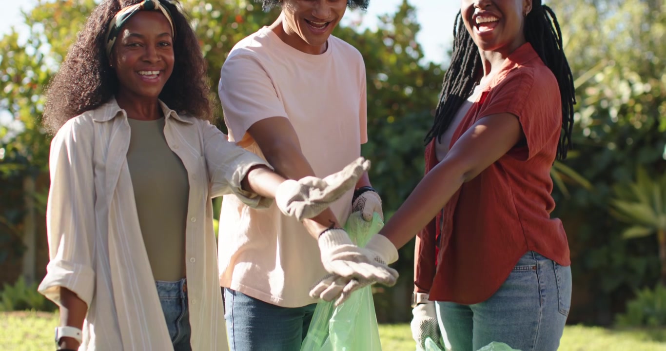 Amigos afro-americanos sorrindo, vestindo luvas cinzas, empilhando as mãos sobre uma sacola verde para limpeza do parque