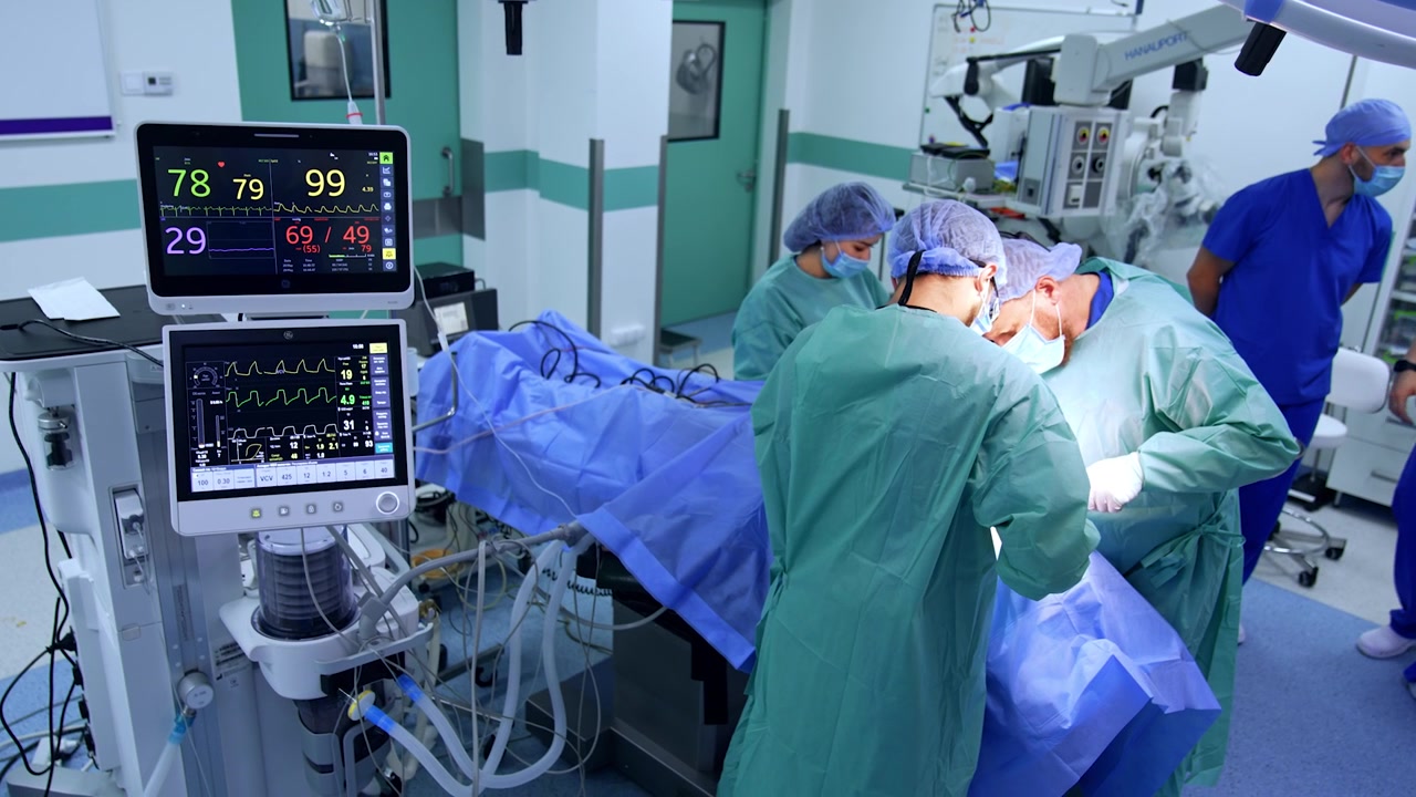 I monitor della macchina per la ventilazione polmonare artificiale mostrano i segni vitali del paziente sottoposto a operazione. Due chirurghi eseguono un intervento di chirurgia maxillo-facciale. Vista dall'alto.