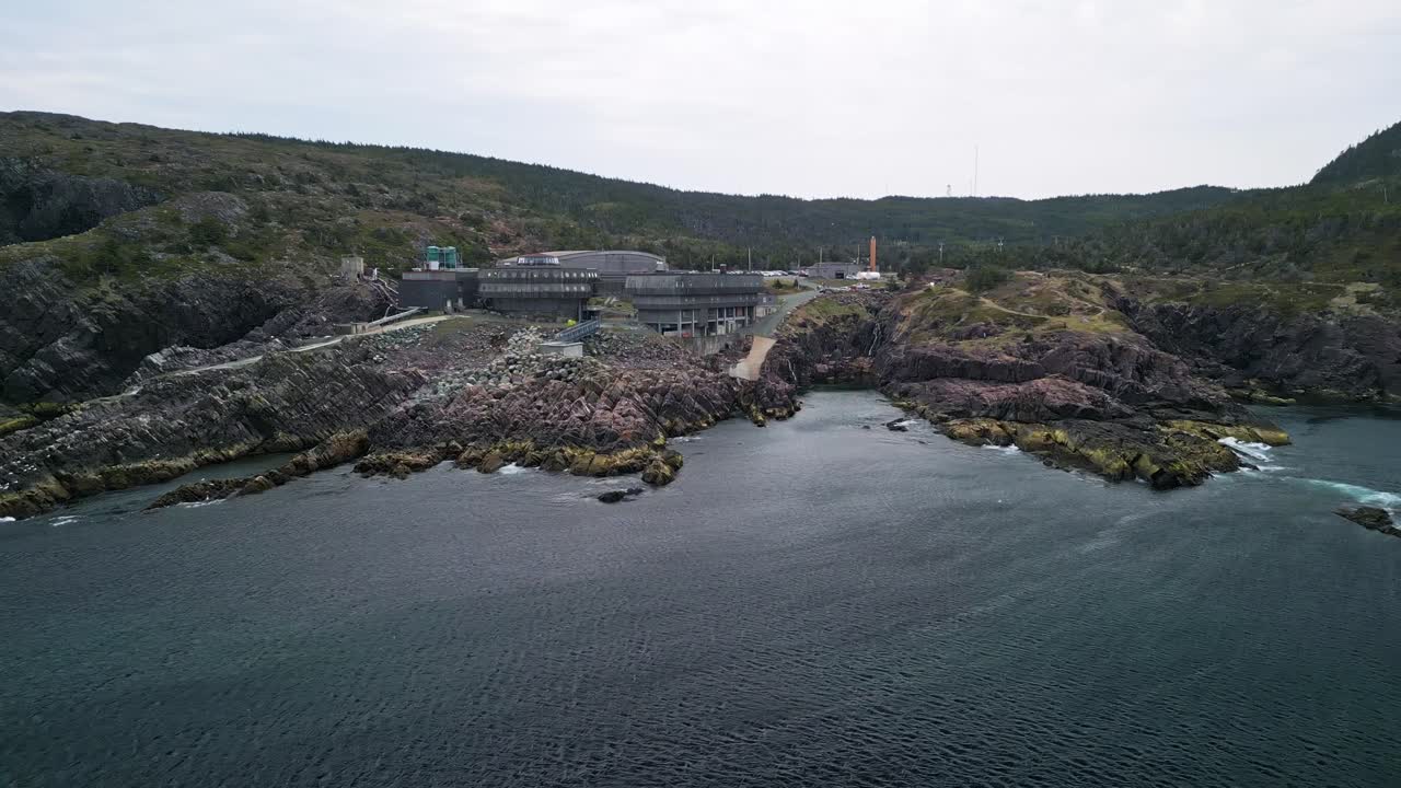 Aerial drone shot circling Logy Bay Marine Lab, Newfoundland. Waves crash below as rocky cliffs and seabirds frame the oceanfront facility.