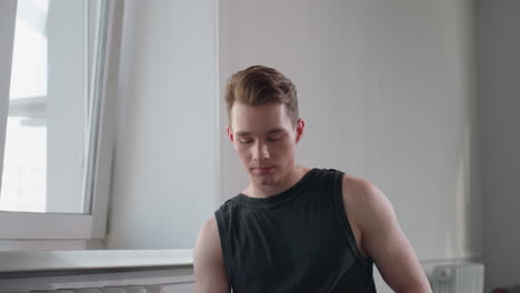 stylish young man with geometric neck tattoo sits indoors by tilted window, wearing black sleeveless top, turning gaze to side in calm thoughtful pose