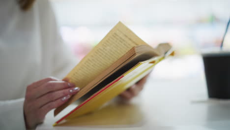 close-up of hands flipping through pages of an open yellow book on a table, with a blurred background featuring a coffee cup, creating a serene and thoughtful atmosphere