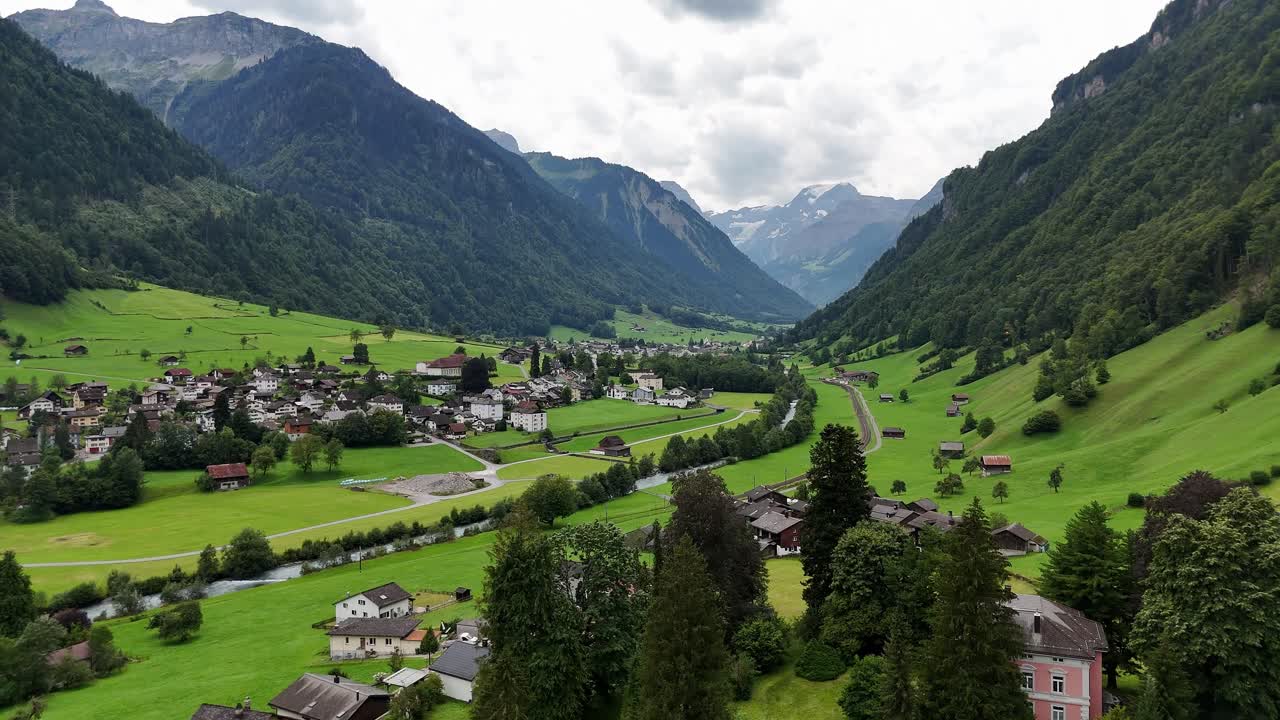 Idyllic landscape in the valley of swiss mountains. Green fields and hilltops with river and small swiss town in summer. Aerial forward wide shot.