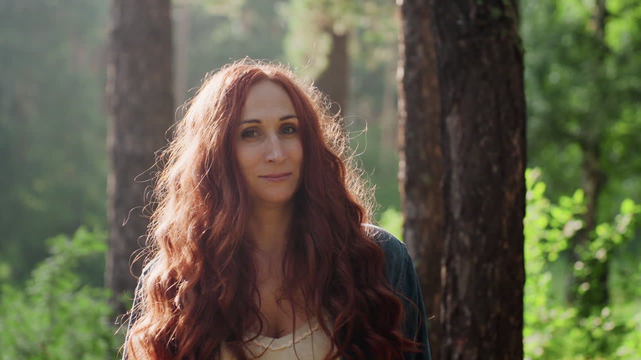 Portrait of picnic lover with long red hair smiling warmly in natural forest, sunlight glowing through trees, relaxed expression showing joy and peace in calm outdoor environment