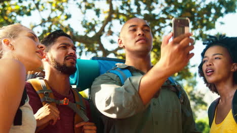 Grupo-De-Amigos-Perdidos-Con-Mochilas-Usando-Gps-En-Teléfonos-Móviles-En-Vacaciones-Caminando-En-El-Campo