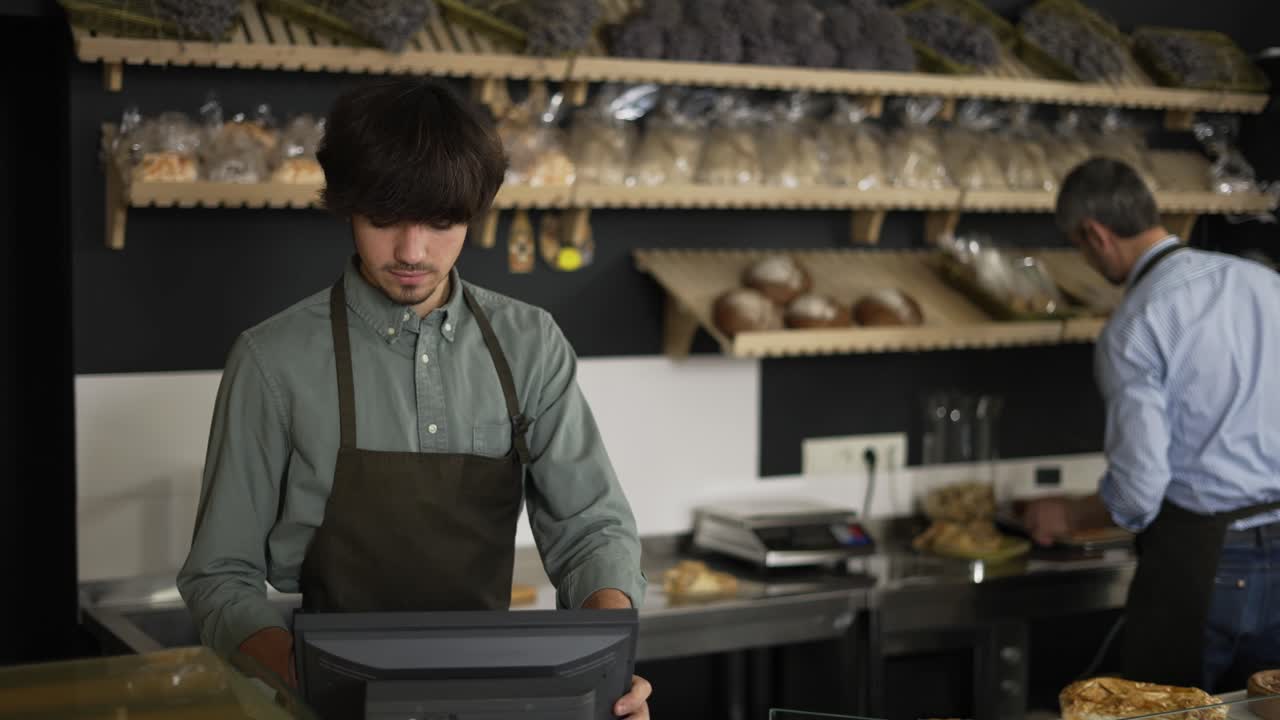 joven cajero en delantal escribiendo en pantalla táctil, guapo trabajador de panadería cortando pan en el fondo