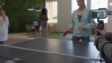 young-business-people-playing-ping-pong-in-office-enjoying-competitive-fun-colleague-using-smartphone-sharing-game-on-social-media