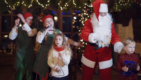 festive atmosphere in santa's residence. santa claus, two elves and children dancing and singing together at christmas.