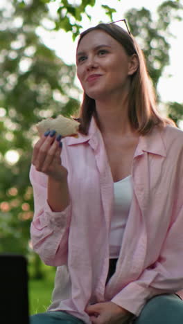 amigos disfrutando de sándwiches a la parrilla al aire libre, una mujer en rosa sentada en la hierba con sándwich en la mano, portátil en el frente, y vegetación en el fondo, disfrutando la comida juntos en un entorno natural