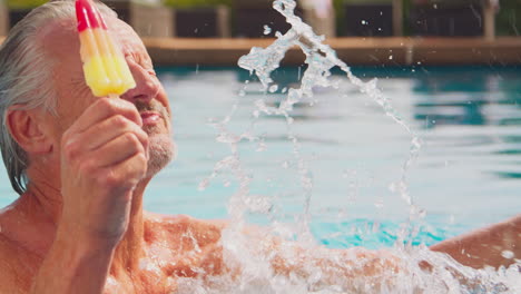 grandson splashing grandfather as they eating ice lollies at edge of swimming pool on summer holiday