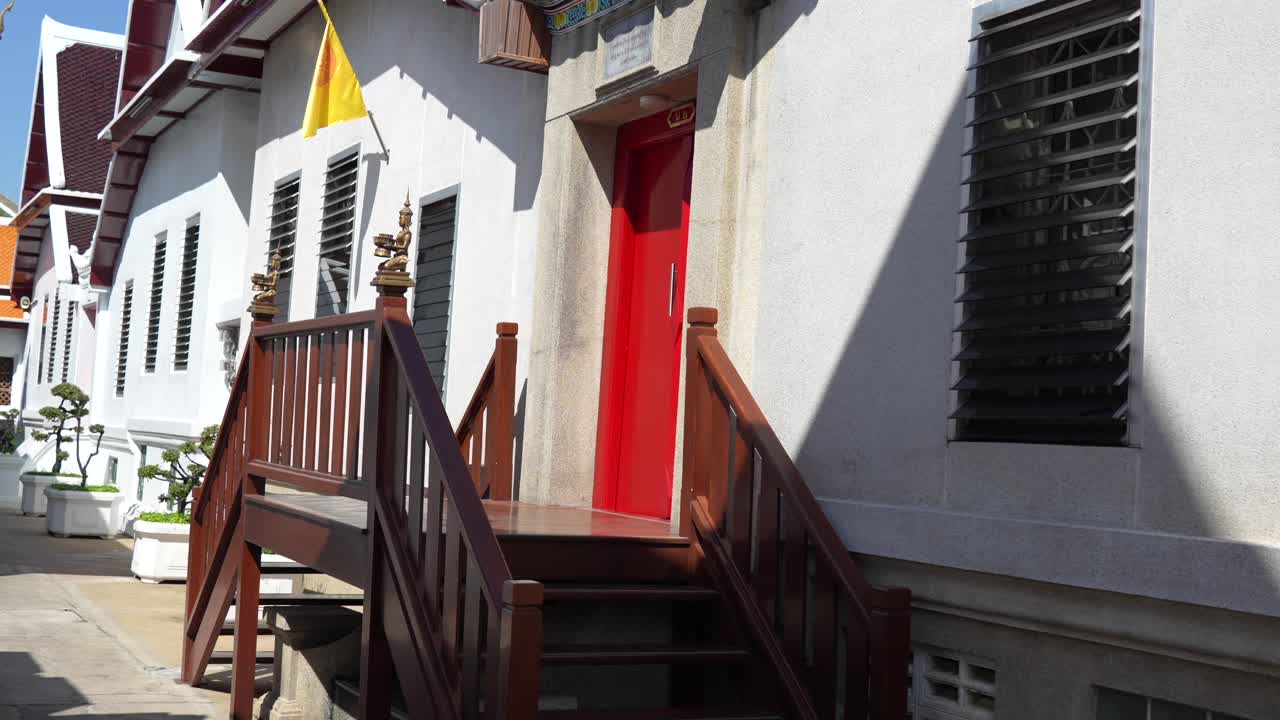 Vista sulla strada a Bangkok, in Thailandia, con un edificio tradizionale caratterizzato da una porta rosso acceso, scale in legno e decorazioni elaborate sul tetto. Un mix di semplicità moderna e dettagli architettonici tailandesi.