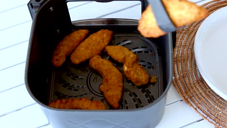 a hand uses tongs to remove crispy chicken from an air fryer onto a plate. bright, clean kitchen setting