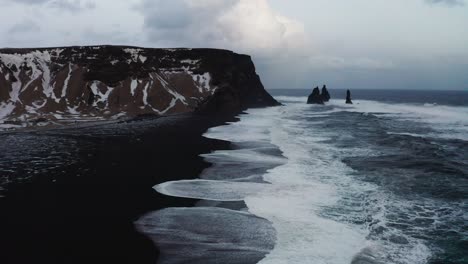 a beautiful drone shot of the black beach in iceland shows the high waves of the sea and the cloudy weather while the snow covers some of the black sand