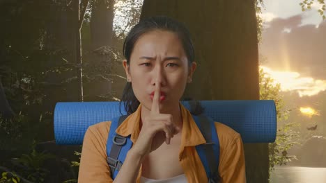 close up of asian female hiker with mountaineering backpack looking at camera and making shh gesture while exploring forest nature