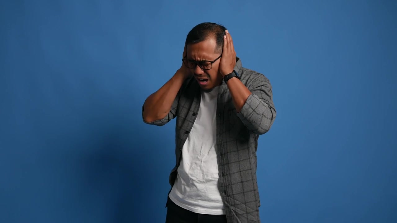 Adult man standing isolated on blue background, stressed covering ears due to loud noise.