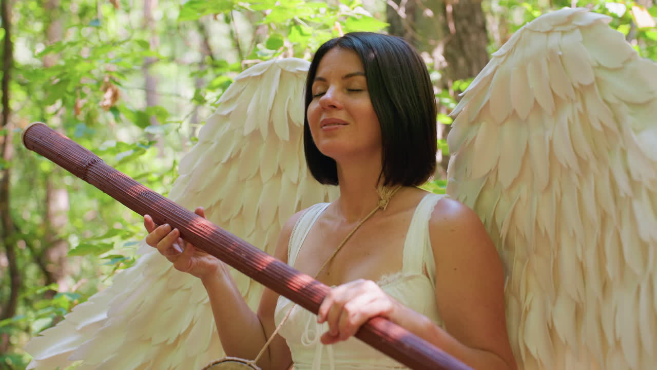 Peaceful angel woman with white wings meditating in green forest holding wooden staff eyes closed surrounded by sunlight and leaves symbolizing inner harmony