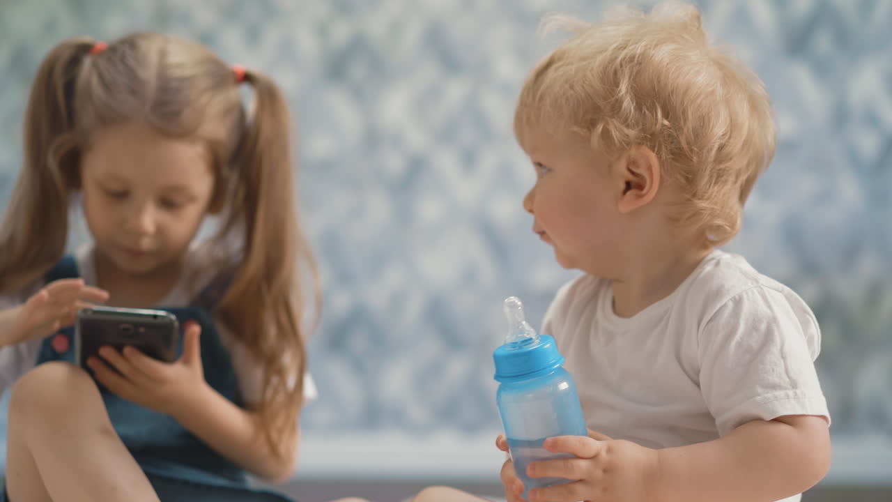 un bambino con una bottiglia si siede vicino alla sorella che gioca con il telefono