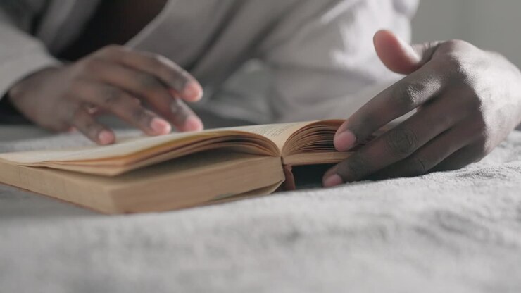 Close-up of hand gently turning pages of large book while lying on bed, focused on reading, soft light filtering through window, creating a peaceful, serene atmosphere in intimate indoor space