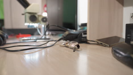 una vista de cerca de una mano derecha con un guante negro alcanzando sus gafas en un escritorio de madera en un entorno de laboratorio, con alambre y otras herramientas electrónicas en la mesa