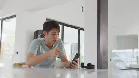 asian boy using smartphone while having breakfast in living room at home