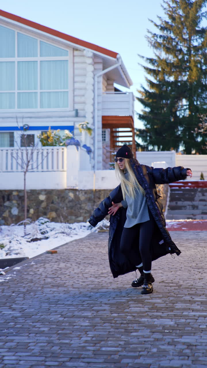 Blonde long-haired woman wearing grey sweater and black leggings puts on a coat outdoors. Girl hops and turns around joyfully. Vertical video.