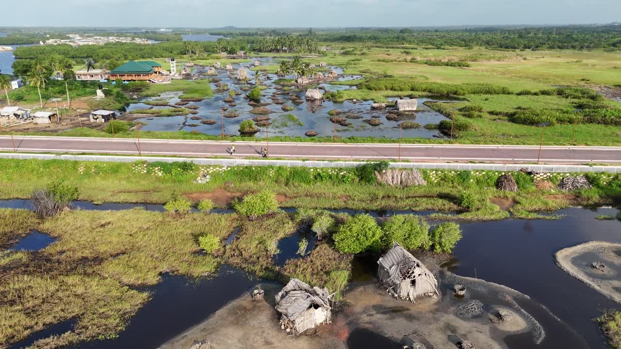 Ripresa aerea da drone dall'alto di una strada moderna che divide le lussureggianti paludi di Ouidah, Benin. Il filmato mostra capanne tradizionali con tetto di paglia costruite sull'acqua in un remoto paesaggio africano.