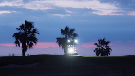un tractor con faros corta la hierba en un campo de golf durante la hora azul de la madrugada, mientras el viento susurra las siluetas de palmeras en el fondo