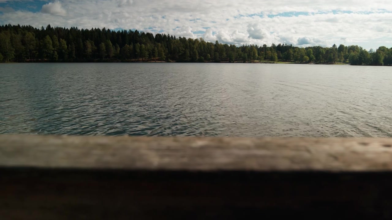 A rough wooden dock rail runs along the bottom of the frame with calm reservoir water and a forested shore under broken clouds in Norway, suited to themes of escape, quiet and reflection.
