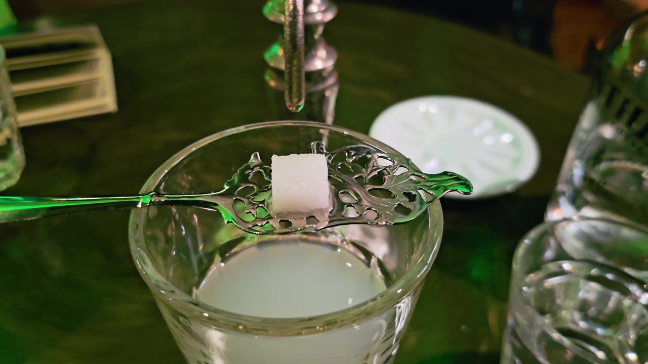 Cận cảnh phục vụ absinthe louche với viên đường trên thìa có rãnh đặt trên ly, giọt nước rơi từ vòi, đồ uống đục, ánh sáng xanh của quán bar và không khí cocktail đầy tâm trạng