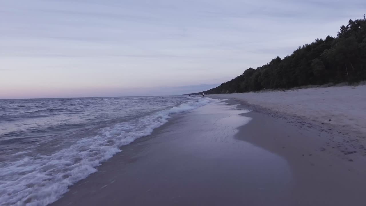 A near-ground drone shot during a sunset on the beach.
You can see the waves on the beach and unrecognizable people taking a walk in the background.