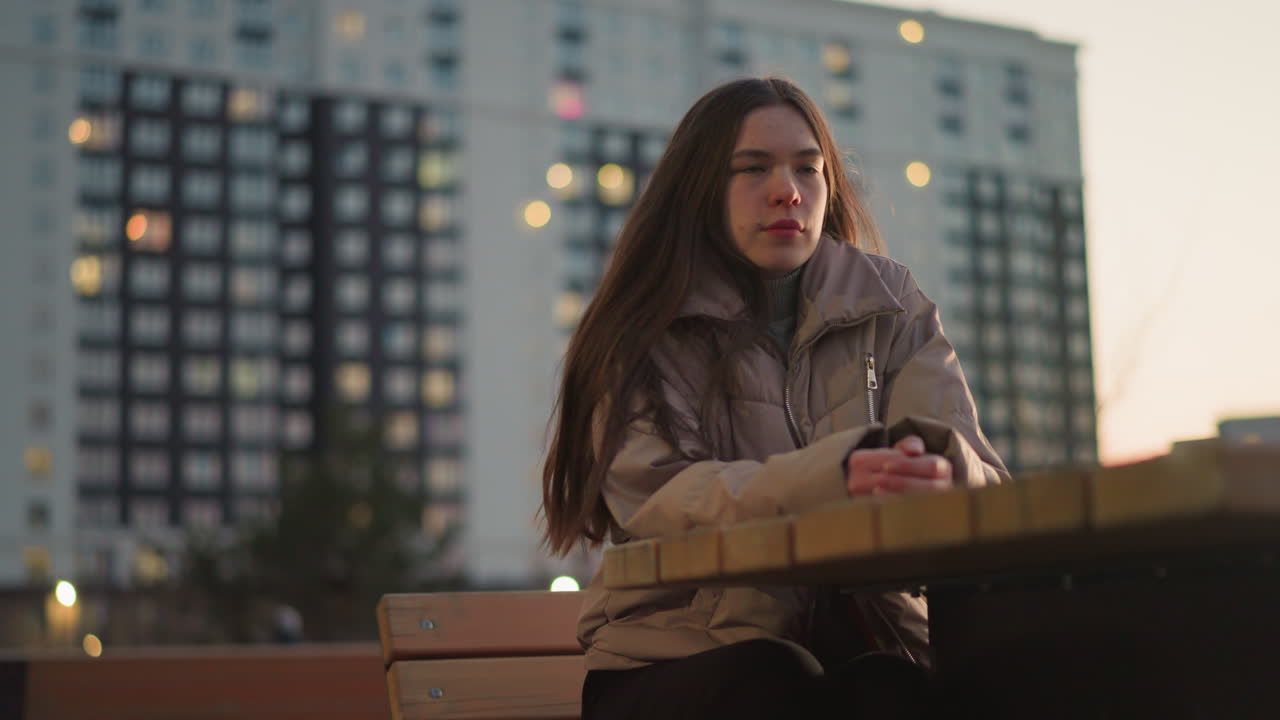 색 재과 검은색 바지를 입은 소녀가 도시 공원의 벤치에 앉아있다. 그녀의 손은 그녀의 앞의 테이블에 휴식을 취하고, 그녀는 그녀의 얼굴에 슬픈 표정을 가지고있다. 저녁 빛과 흐릿한 배경