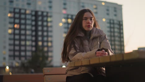 una chica con una chaqueta de melocotón y pantalones negros está sentada en un banco en un parque urbano. sus manos descansan en la mesa frente a ella, y tiene una mirada triste en su rostro. luz de la noche y fondo borroso