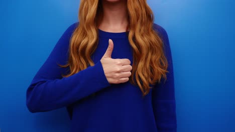 Joven-Pelirroja-Con-Camisa-Azul-Mostrando-Un-Gesto-De-Pulgar-Hacia-Arriba,-Aislada-En-El-Fondo-Azul