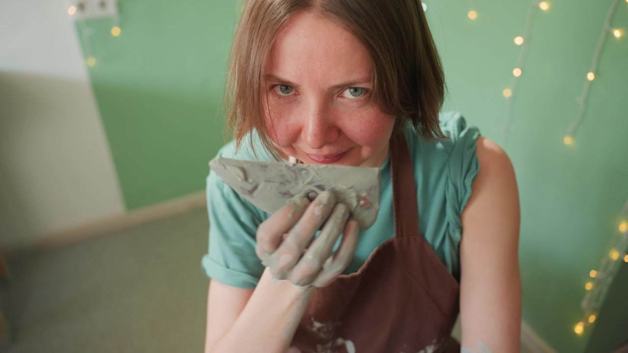 Happy artist shaping small ceramic cup with muddy hands and cheerful expression. Enthusiastic female creator enjoying relaxed pottery session with luminous lighting and positive vibe
