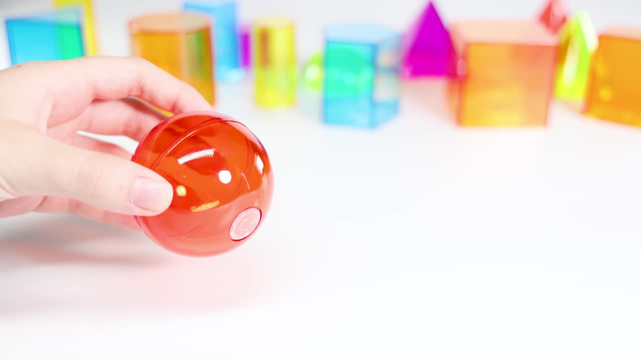 A hand interacts with a red sphere among colorful 3D shapes on a white surface, emphasizing geometry and color