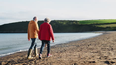 Rückansicht-Eines-Aktiven-älteren-Paares,-Das-Sich-An-Den-Händen-Hält-Und-Im-Winterurlaub-Am-Strand-Entlang-Der-Küste-Spaziert