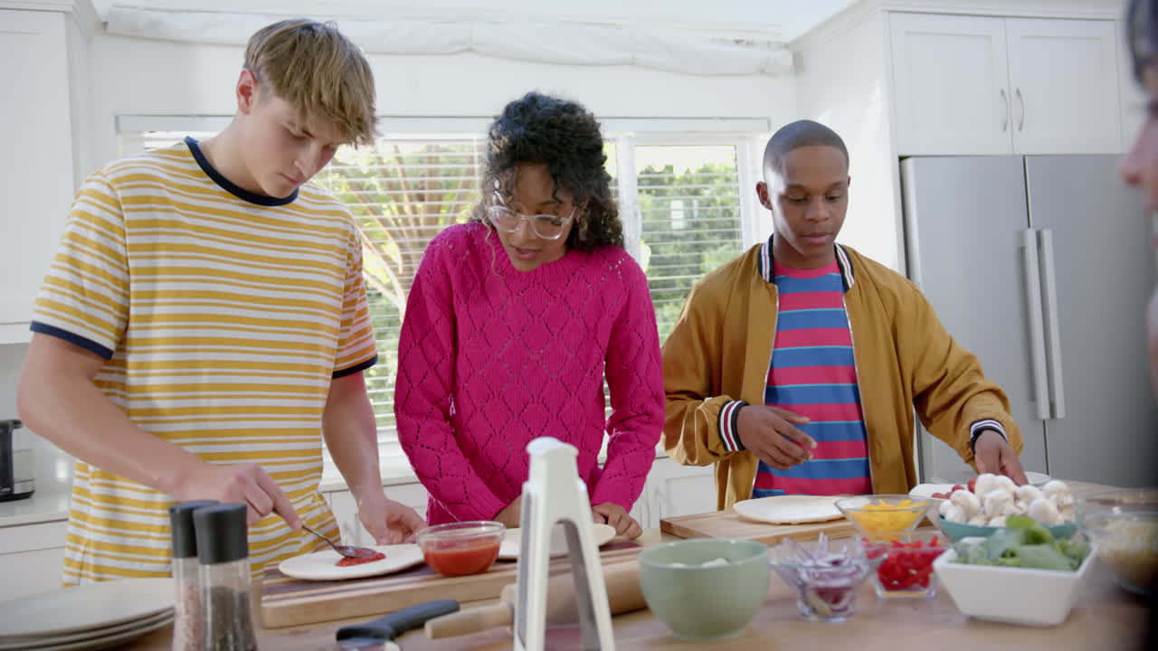 Happy diverse group of teenage friends cooking and making pizza in kitchen, slow motion