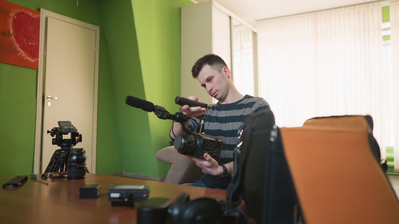 Man reaching into camera equipment bag for microphone in bright, casual workspace. Camera gear such as lenses and tripod on table. Green walls and natural lighting create cozy atmosphere