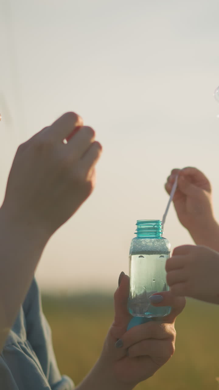 A close-up of a woman in a blue dress kneeling in a grassy field at sunset, holding a bubble bottle as she and her young son, dressed in a white shirt, blow bubbles from wands
