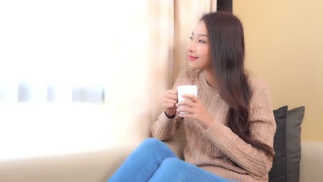 young satisfied asian woman drinking coffee or team by window ay sofa in living room of family home, static full frame slow motion