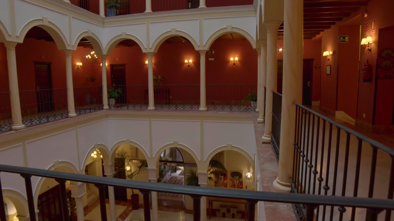 A wide shot panning left showcases the elegant courtyard of a Spanish hotel, featuring arched walkways, warm lighting, and classic decor with plants and chandeliers.